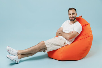 Sticker - Full length young smiling friendly happy man 20s in casual white t-shirt sit in bag chair hold hands crossed folded rest relax isolated on plain pastel light blue color background studio portrait