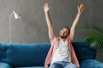 Poster - Young smiling happy fun satisfied relaxed man 20s wear casual clothes do morning hand stretching sitting on blue sofa at home flat indoors rest on weekends free time. People lounge lifestyle concept