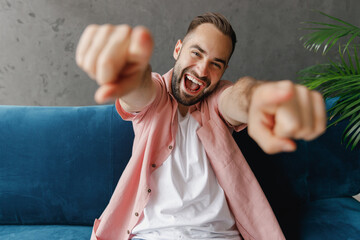 Wall Mural - Young smiling happy fun confident man 20s in casual clothes point index finger camera on you sitting on blue sofa at home flat indoors rest relax on weekends free time People lounge lifestyle concept