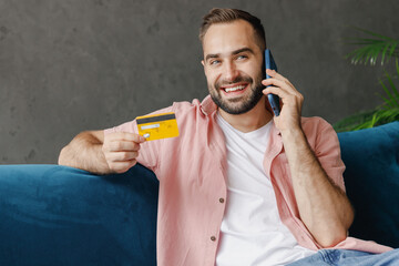 Wall Mural - Young cool smiling man 20s in casual clothes talking speak on mobile cell phone hold credit bank card booking delivery shopping online sit on blue sofa at home flat indoors rest relax on weekends.