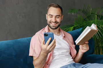 Wall Mural - Young smiling happy man 20s wear casual clothes reading book novel use mobile cell phone sitting on blue sofa at home flat indoors rest relax on weekends free time. People lounge lifestyle concept