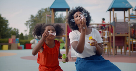 Afro-american mother and daughter play soap bubbles at playground.