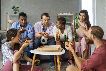 Casual group of happy young diverse friends relaxing on floor and sofa, eating pizza slices from carton box ordered in delivery, drinking beer, discussing sports games on TV, having good time together