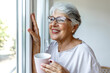 70s grandmother start new day with cup of hot coffee or tea, favourite beverage, standing in cozy living room looking out the window. Senior woman daydreaming enjoy calm retired life concept.