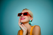 Happy Smiling Fashionable Woman Wearing Trendy Summer Orange Sunglasses, Green Shell Earrings, Posing On Blue Background. Model Looking Up. Copy, Empty Space For Text
