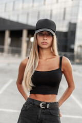 Fashion pretty young woman with a beautiful athletic body in a black T-shirt with a black cap for design mockup in black jeans stands in the city