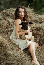Brunette Caucasian Woman Sitting On The Hay Grass Holding A Baby Goat Wearing A White Dress