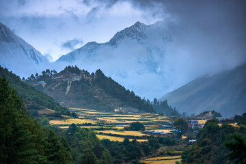 Wall Mural - Small village on the mountain peak in fog at sunset in Nepal. Colorful landscape with houses, blue sky with low clouds, high rocks, green forest and yellow fields at dusk. Travel in Himalayas. Nature