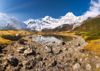 Wall Mural - Manaslu mountain and lake at sunrise in Nepal. Beautiful landscape with snowy mountains, stones, mountain lake, reflection, grass, blue sky and sun beams. Rocks in snow. Himalayan mountains. Himalayas