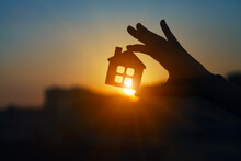 Man Holding  Wooden House Model Against Sunset Light For Sale Or Rent, Family Home And Shelter Concept, Real Estate, Solar Energy And Eco Accommodation