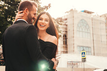 Beautiful Fashion Woman And Her Handsome Boyfriend In Suit. Sexy Smiling Brunette Model In Black Evening Dress. Fashionable Couple Posing In The Street At Sunset. Brutal Man And His Female Outdoors