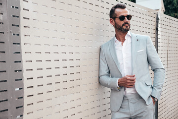 Portrait of handsome confident stylish hipster lambersexual model.Modern man dressed in elegant white suit. Fashion male posing in the street background near fence at sunset. In sunglasses