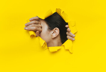 Wall Mural - Close-up of a woman's ear and hands through a torn hole in the paper. Yellow background, copy space. The concept of eavesdropping, espionage, gossip and tabloids.