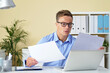 © DragonImages - Serious young businessman in glasses reading contracts and reports at his office desk