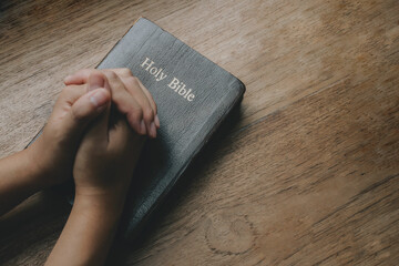 Woman with Bible praying, hands clasped together on her Bible on wooden table.