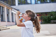 Schoolgirl girl drinks fresh mineral water from a bottle in the schoolyard. Take a break from school. Quenching thirst, replenishing water balance in children