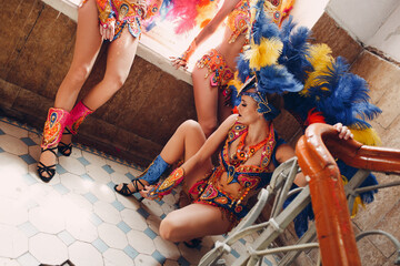 Wall Mural - Woman in brazilian samba carnival costume with colorful feathers plumage relax in old entrance with big window.