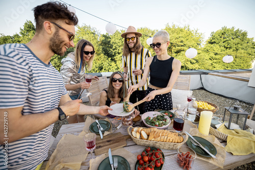 Young stylish friends have a festive dinner outdoors, share healthy food, have great summer time together on a rooftop terrace