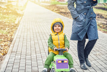 Happy Little Boy Kid In Bright Yellow Vest Pushes Ride-on Toy Walking With Mother Wearing Grey Coat Along Road In Autumn Park