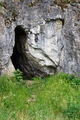 Canvas Print - The entrance to the cave in the rock with grass in the foreground.