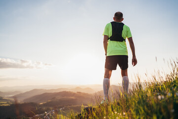 Wall Mural - Backview of Active mountain trail runner dressed bright t-shirt with backpack enjoying the sunset while he sky running by picturesque hills at sunset time. Sporty active people concept image