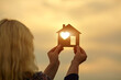 Woman holding a wooden house in front of the sun