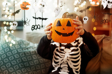 Wall Mural - halloween, holiday and childhood concept - smiling little boy in costume of skeleton with jack-o-lantern sitting on sofa at home