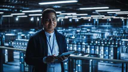 Handsome Smiling IT Specialist Using Tablet Computer in Data Center, Looking at Camera. Succesful Businessman and e-Business Entrepreneur Overlooking Server Farm Cloud Computing Facility.