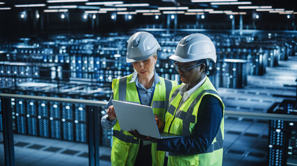 Leinwandbilder - Data Center IT Specialist and System administrator Talk, Use Laptop Computer, Wearing Safety West. Server Cloud Farm Facility with Two Female Information Technology Engineers Doing Maintenance.