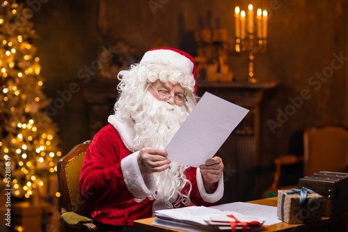 Workplace of Santa Claus. Cheerful Santa is reading letters from children while sitting at the table. Fireplace and Christmas Tree in the background. Christmas concept.