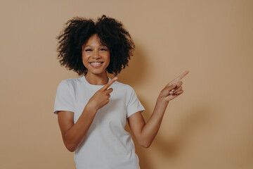 Wall Mural - Positive cheerful dark skinned woman pointing at copy space isolated on beige background