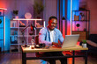 © sofiko14 - Handsome african man in casual clothes sitting at table and typing on wireless laptop. Young guy doing remote work on portable computer during evening time at home. Freelance concept.