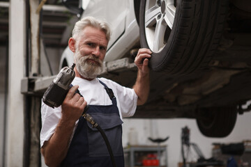 Wall Mural - Elderly bearded car service worker holding torque wrench, standing near lifted car, copy space