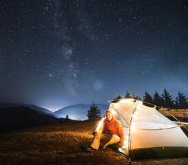 Wall Mural - Young traveler sitting in his white tent, lit by light bulbs, on grass under open starry sky with Milky way. Night mountain camping against backdrop of mountain hills and light of city between them.