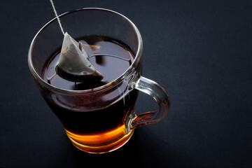 Wall Mural - Black tea in a pyramid tea bag is brewed in hot water in a transparent cup. Black background. Diagonal top view. Free space for an inscription