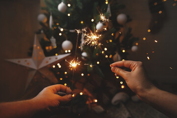 Wall Mural - Hands holding burning sparklers on background of christmas tree and glowing star in festive scandinavian room. Happy New Year! Couple celebrating with firework bengal lights. Atmospheric moment