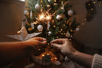 Poster - Happy New Year! Couple celebrating with firework bengal lights on background of christmas tree and glowing star. Hands holding burning sparklers in festive scandinavian room. Atmospheric moment