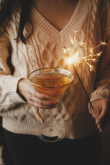 Wall Mural - Woman holding burning sparkler and champagne glass, celebrating and toasting at party. Happy New Year! Firework bengal light and drink in woman hands. Atmospheric moment. ​Happy Holiday