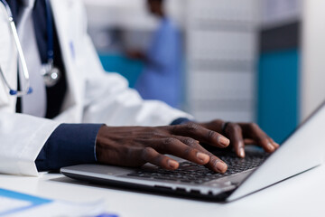 Wall Mural - Close up of black hands typing on laptop keyboard while sitting at white desk in medical office. African american man wearing medical coat using computer device and modern technology