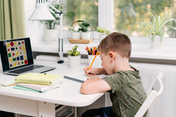 Wall Mural - Distance learning online education. Caucasian smile kid boy studying at home with laptop and doing school homework. Thinking child siting with notebook, pencils and training books. Back to school.