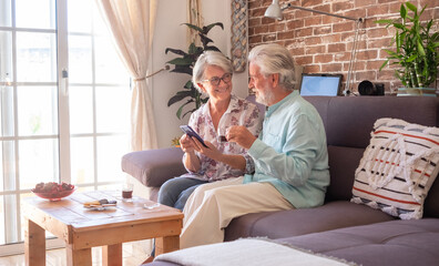 Smiling senior couple at home sitting on sofa with a coffee using mobile phone. Brick wall on background