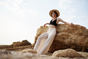 Portrait of woman in black bralette and white trousers leaning on rock at beach