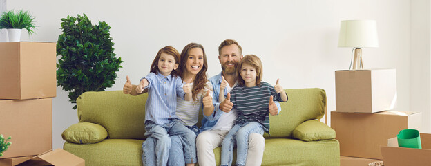 Portrait of smiling family in new home. Happy content mom, dad and kids showing thumbs up gesture sitting on sofa in room with unpacked boxes. Real estate, mortgage, buying property, ownership concept