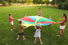 Group Of Children And Teachers Playing With Rainbow Playground Parachute On Green Grass. Summer Camp Activity