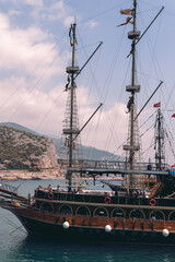 Wall Mural - Vertical photo. Old pirate ship on the water of Mediteranean sea. Tourist entertainment, coastal tour. Summer sunny day. Mountain shore of Alanya. Turkey.