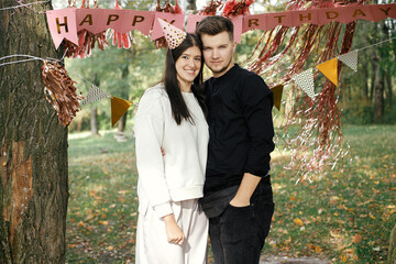 Wall Mural - Happy woman in party hat and handsome man hugging on background of pink happy birthday garland in park. Young stylish couple celebrating birthday with family and friends at picnic party outdoor
