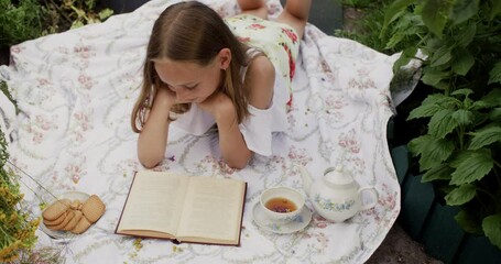 young girl reading book lying on summer meadow with biscuits and tea on carpet. romantic woman with 