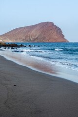 Poster - Vertical shot of a blue seascape with a mountain under a clear sky in Granadilla de Abona