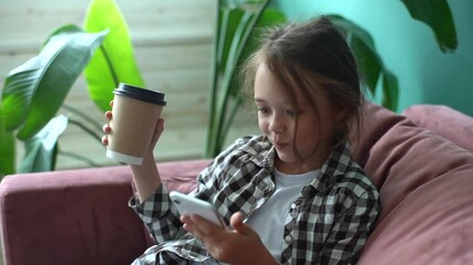 Close-up of amazed little child girl scrolling phone screen and saying wow sitting in armchair holding coffee cup at home. Pretty young girl with long blond hair using smartphone and looking surprised