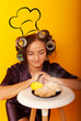 © Marina Chernivetskay - a girl in curlers looks at croissans a housewife cooked homemade cakes on a yellow isolated background